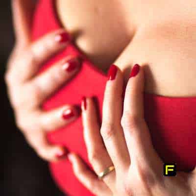 Close-up of a woman's hands gently lifting her bust while wearing a red dress and matching red nail polish. This image represents confidence, self-care, and natural bust enhancement, complementing the article on firming lotions and oils.