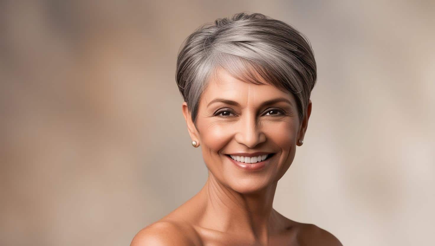 A confident woman over 50 with a stylish, short pixie cut. Her hair is neatly trimmed around the ears with soft, feathered layers on top for volume. She has natural gray hair and a warm, smiling expression. The background is soft and blurred, creating a professional studio portrait. The Best Hairstyles for Women Over 50: 40+ Flattering Cuts & Styles