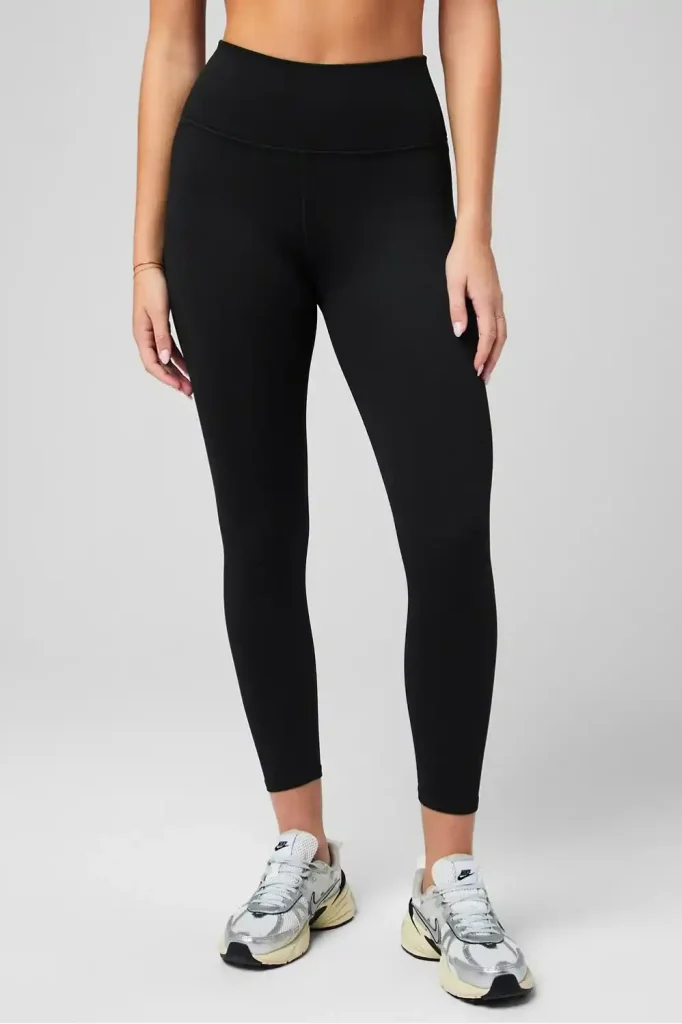 16 Best Workout Leggings for Plus Size Women Over 50 (2025) 9 Woman wearing black high-waisted 7/8 length leggings with a wide waistband. The athletic leggings have a slim fit and end just above the ankles. The model is shown from waist to feet, wearing white and beige Nike athletic shoes against a light gray background.