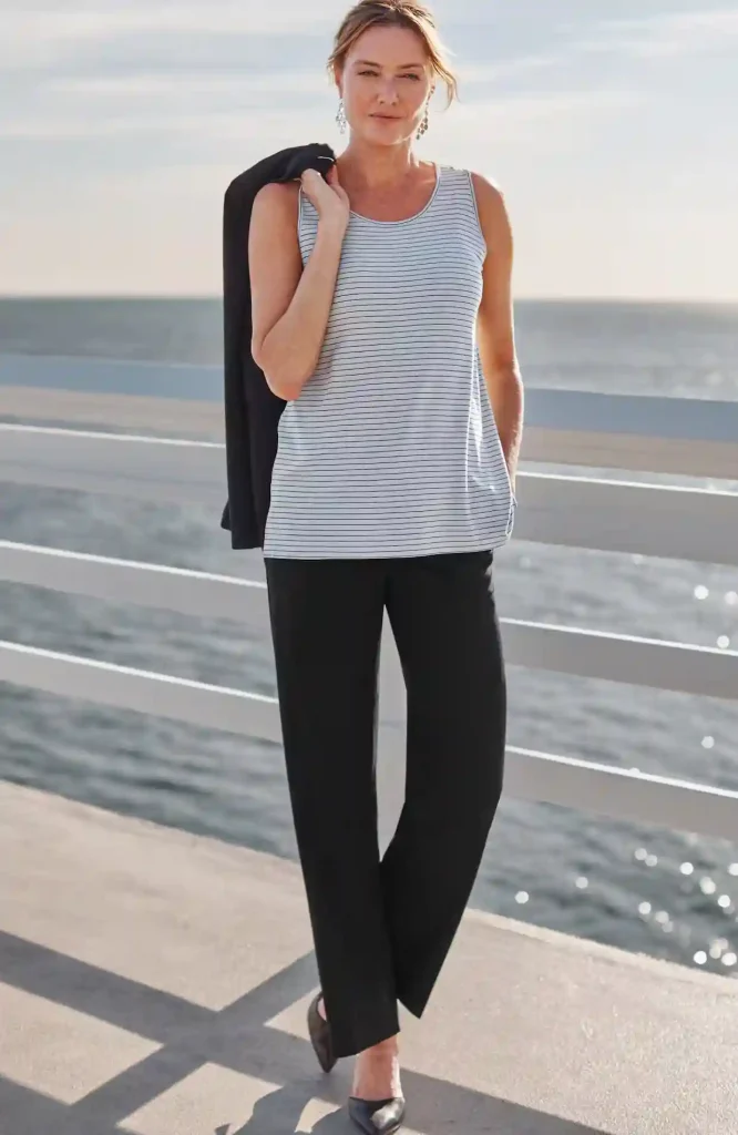 Best Wide Leg Pants for Women Over 50: Stylish, Comfortable, Flattering Options for 2025 9 Woman standing on a boardwalk by the ocean wearing black wide leg pants from the J.Jill Wearever Collection, a striped sleeveless top, and holding a black jacket over her shoulder.