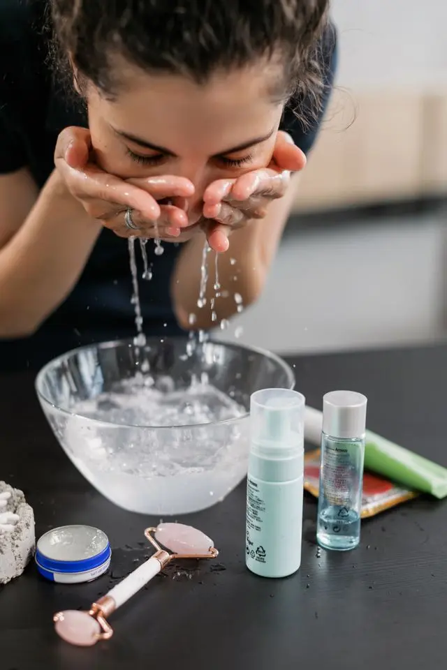 The Best Premium Face Wash for Women with Combination Skin (11 Cult-Favorite Picks) 5 Woman rinsing her face over a bowl with a combination skin cleansing routine and rose quartz roller on the counter.